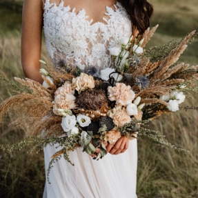 A slender bride in a white lace dress, holding a bridal bouquet of flowers including sprays of wheat ears, ferns and grasses